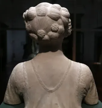 The back of a marble bust of a young woman with an elaborate coiled hairstyle and decorated drapery over the shoulders.