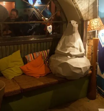 Homunculus Loxodontus sculpture seated on a padded bench among colorful pillows.