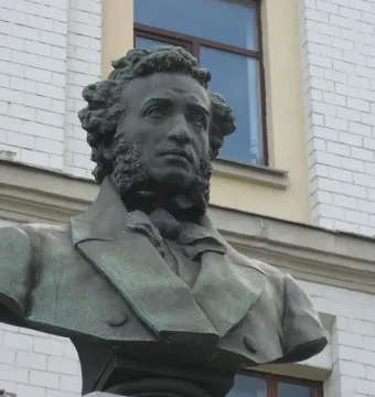 Green-patinated bronze bust of Alexander Pushkin outdoors in front of a light-colored building.