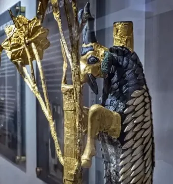 Ram in a Thicket sculpture with a gold head and legs, dark curved horns, and a blue-black and gold body leaning against a tree-like structure.