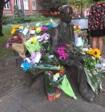Alan Turing Memorial statue on a park bench surrounded by colorful flower bouquets.
