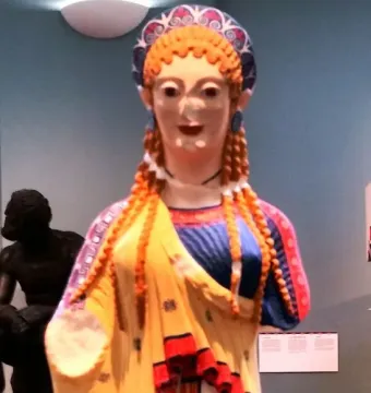 Korè 675, a brightly painted replica of an ancient girl statue with a missing lower arm, standing against a blue museum wall.