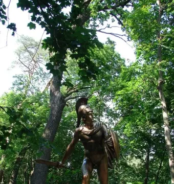 Greek Warrior Monument showing a bronze-toned armored figure with a helmet, sword, and shield among trees.