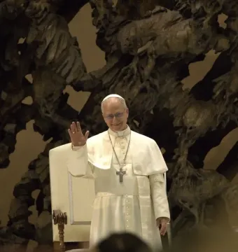 Pope Leo XIV in white clerical vestments stands in front of The Resurrection sculpture, smiling and waving.