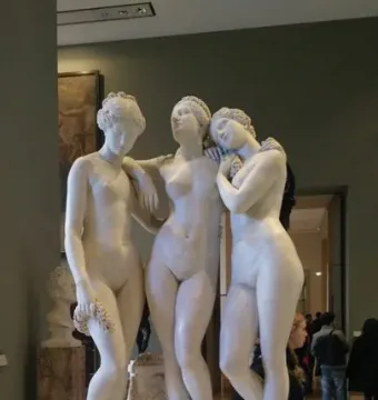 The Three Graces marble sculpture on a tall pedestal in a museum gallery, with blurred visitors in the background.