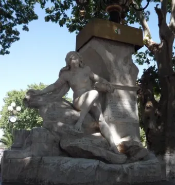 Ain El Fouara Fountain with a reclining nude female statue on a rocky base, framed by trees and a lamppost.