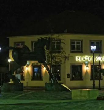 Anscavallo, a dark equine sculpture, stands in a town square at night near a building with an illuminated Platengarten sign.