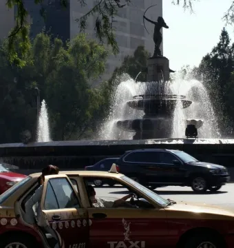 Diana the Huntress Fountain rises above city traffic, with a red and yellow taxi in the foreground.