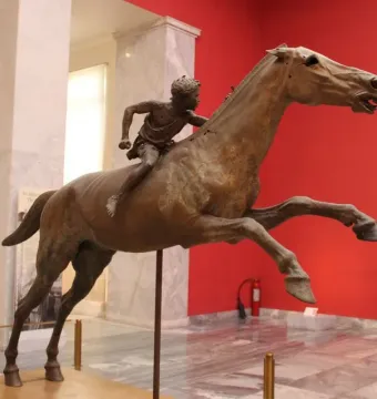 Jockey of Artemision bronze sculpture showing a young rider leaning forward on a horse in motion inside a museum.