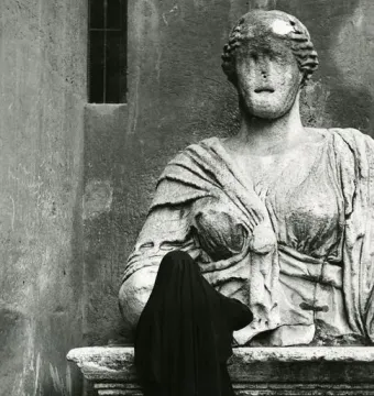 Madama Lucrezia, a weathered stone statue of a woman, stands on a pedestal beside a dark-robed figure seen from behind.