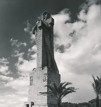 The Monument to Columbus rises on a tiered base beneath a cloudy sky, with palm trees and distant water in the background.