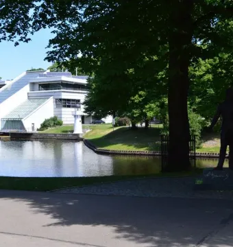 Pushkin statue on a pedestal beneath a large tree, with water and a modern building in the background.