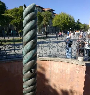 The Serpent Column with a weathered green patina stands inside a shallow red-walled enclosure near an ornate fence and public square.