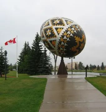 Vegreville Pysanka egg sculpture with geometric gold, black, and silver patterns in a park with trees and a pond nearby.