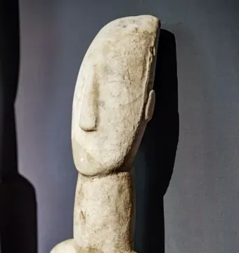 Kykladic Idol marble figurine shown from the upper chest up, with a long neck and distinct nose against a dark background.