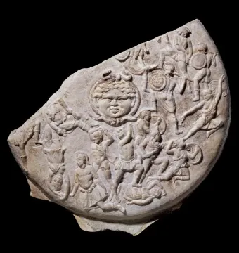 Stone relief fragment showing a central face with serpentine hair surrounded by densely packed warriors in battle.