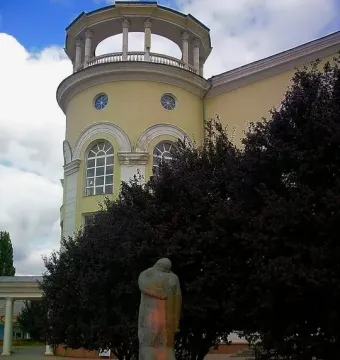 The Shot in the Back monument showing two embracing figures on a dark base with a wreath, set among trees with a towered building behind it.