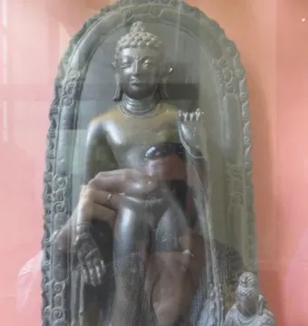 Sultanganj Buddha displayed in a glass case with a kneeling figure beside it.