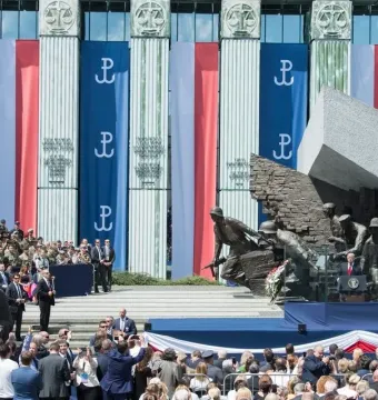 Warsaw Uprising Monument with a speaker addressing a crowd in front of it and Polish banners on a nearby building.