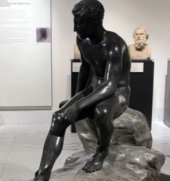 Seated Hermes statue of a nude male figure with winged sandals, shown on a rocky base in a museum display.