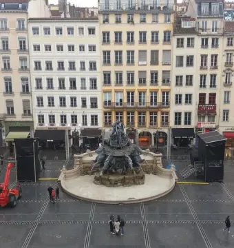 Fontaine Bartholdi in the center of Place des Terreaux, surrounded by wet paving, pedestrians, and surrounding buildings.