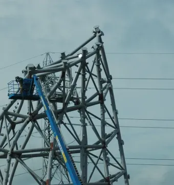 A large grey metal sculpture under construction with a worker in a blue boom lift nearby and power lines overhead.