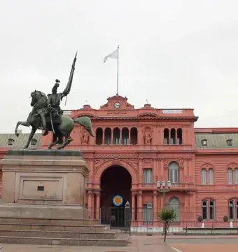 Manuel Belgrano equestrian monument with a rearing horse and raised standard in front of a pink government building.