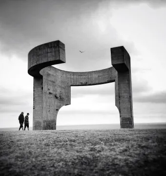 Elogio del Horizonte concrete sculpture against a cloudy sky, with two people walking on the grass below and the ocean in the background.