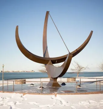 Man Enters the Cosmos sundial sculpture on a circular base beside a snowy lakeshore, with a city skyline in the distance.