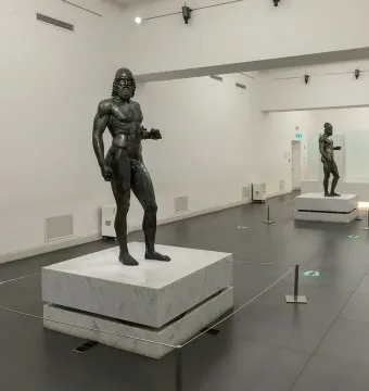The Riace bronzes displayed on white pedestals in a brightly lit museum hall.