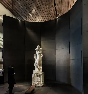 Michelangelo’s Rondanini Pietà displayed on a pedestal in a dim museum gallery, with a visitor standing nearby.