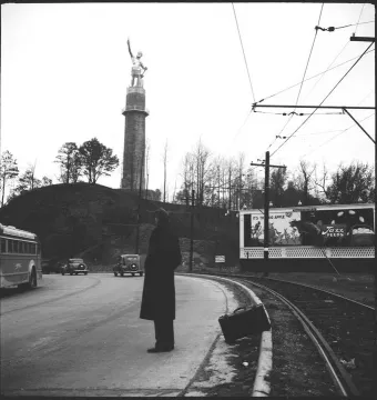 A distant Vulcan statue rises on a hill above a city street with railroad tracks, vintage cars, and a bus in the foreground.