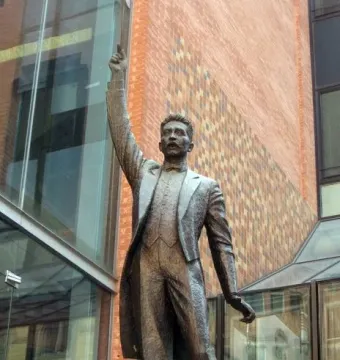 A bronze statue of A Lluís Millet in a formal suit and bow tie, with one arm raised and pointing upward.