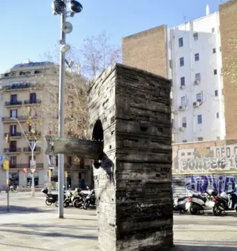 Monument to Narcís Monturiol, a tall textured concrete block with a submarine-like metal model protruding from its side, on a city sidewalk.