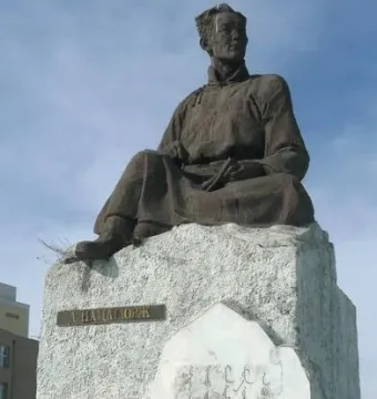 Natsagdorj Monument showing a weathered bronze-colored seated figure on a stone pedestal with engraved inscriptions and a floral wreath at the base.
