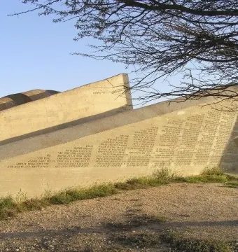The Negev Brigade Monument, a large angular concrete structure covered with inscriptions, stands on rocky ground beneath a clear blue sky.