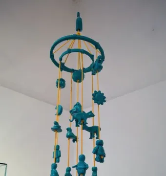 Teal cartonería mobile with small figures hanging from yellow strings, including animals, celestial shapes, and abstract forms.