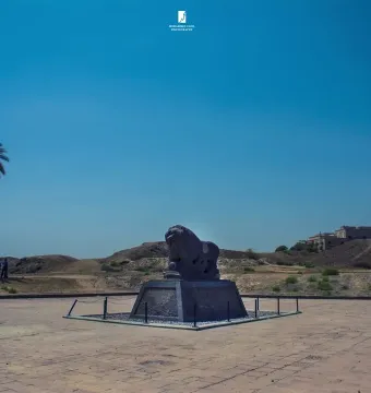 Lion of Babylon sculpture on a modern pedestal in a paved area with palm trees and distant hills behind it.