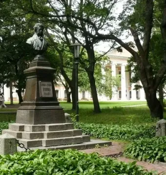 Bust of Pushkin on a tall stone pedestal in a park, with trees and a columned building in the background.
