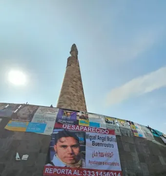 The Roundabout of the Disappeared monument rising against a bright blue sky, with posters and a disappeared-person flyer covering its base.