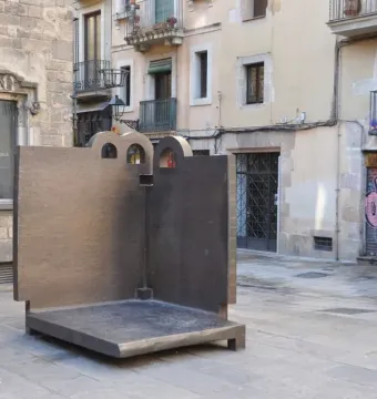 Topos V, a large dark L-shaped sculpture with arched cut-outs, standing on a stone-paved plaza in Barcelona.