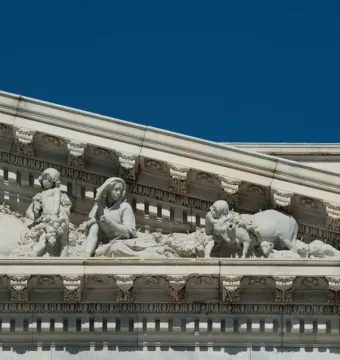 Apotheosis of Democracy pediment relief showing carved figures with livestock, a child carrying grapes, and a seated woman under a blue sky.