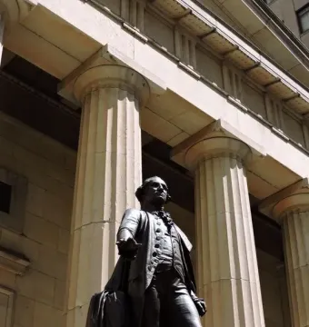 The Statue of George Washington stands on a stone platform before a classical building with tall fluted columns.