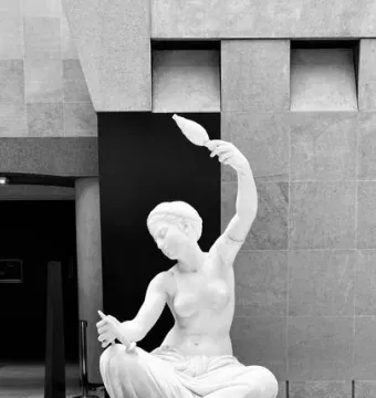 Jeune fille de Mégare, a white marble statue of a seated draped woman holding an object aloft in her raised hand.