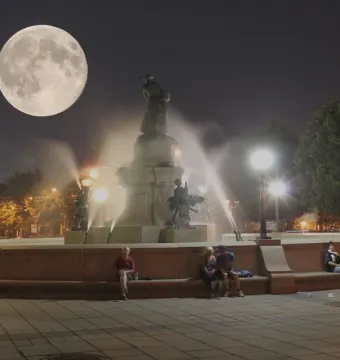 La Fermière fountain in Montreal lit at night with water jets and a full moon overhead.