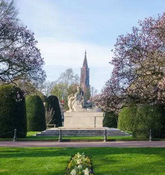 Stone war memorial sculpture with several figures on a stepped base, framed by pink-blossomed trees and trimmed hedges.
