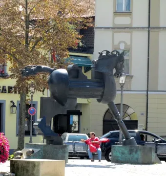 Anscavallo, a dark abstract horse monument in a town square beside flower beds and buildings.