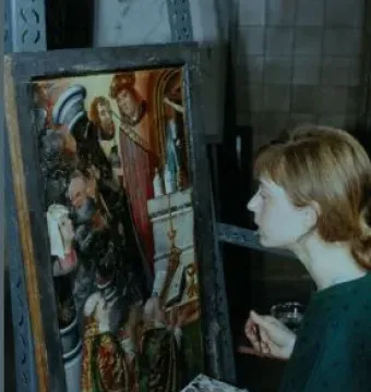 A conservator examines the Fisenne Altarpiece while holding a brush and palette beside the painted carved panel.