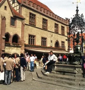 Gänseliesel Fountain in a town square, with a statue beneath an ornate dark spire and people sitting on the steps.