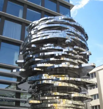 The Head of Franz Kafka sculpture with stacked, mirrored metal panels reflecting nearby buildings and blue sky.